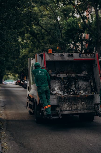 A waste collection worker wearing a green uniform and helmet stands next to an open, rear-loading waste collection truck on a residential street lined with trees and parked cars. The truck is primaril