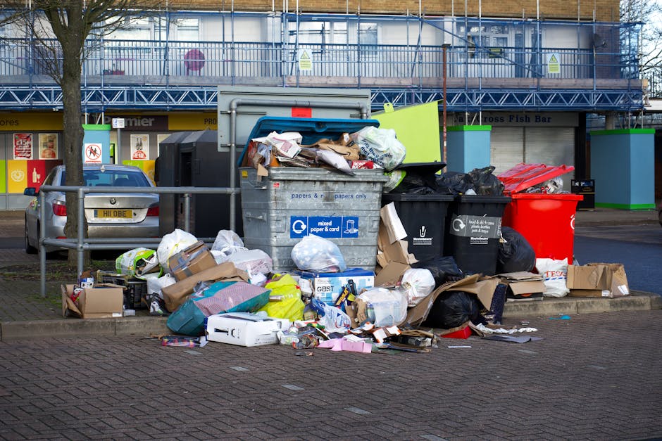 A collection of overflowing rubbish and waste materials accumulated on a paved city street near a parking area. The pile includes various types of waste such as crumpled paper, cardboard boxes, plasti