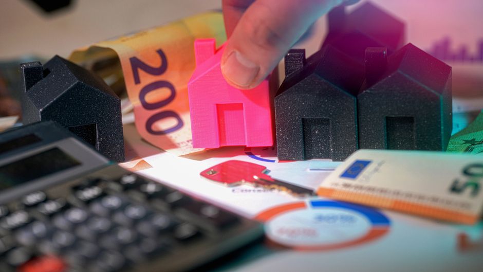 A close-up view of a person's hand placing a small, bright pink, house-shaped model among several black house-shaped models with textured surfaces on a cluttered surface. Surrounding the models are va