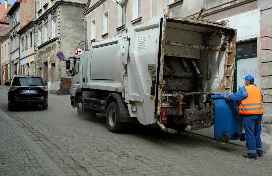 A large collection of rubbish and mixed waste is piled in and around multiple wheelie bins on a paved urban sidewalk. The waste includes crumpled cardboard boxes, plastic bags, paper, and packaging ma